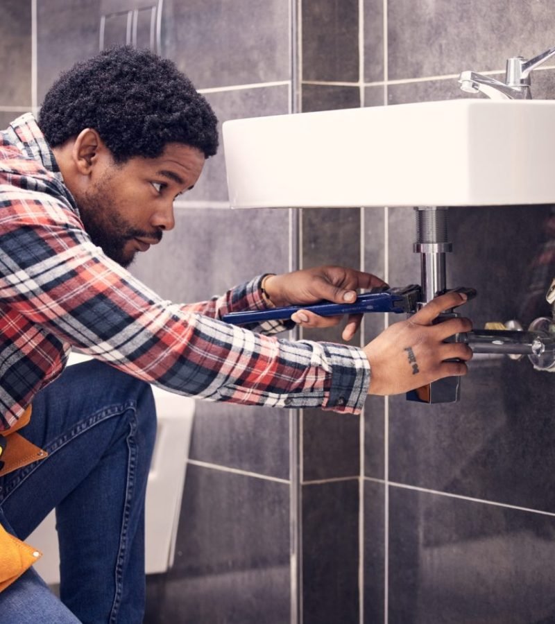 black-man-plumber-and-maintenance-in-bathroom-fixing-sink-pipe-with-wrench-and-plumbing-industry--e1705240801490
